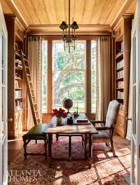 Wood-paneled study with built-in bookshelves, a rolling ladder, a large wooden desk with a globe and flowers, and a window overlooking a large tree.