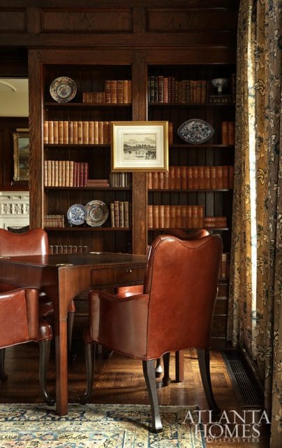 Dark wood paneled library with floor-to-ceiling bookshelves filled with leather-bound books, a framed landscape painting, decorative plates, and brown leather chairs around a wooden table.
