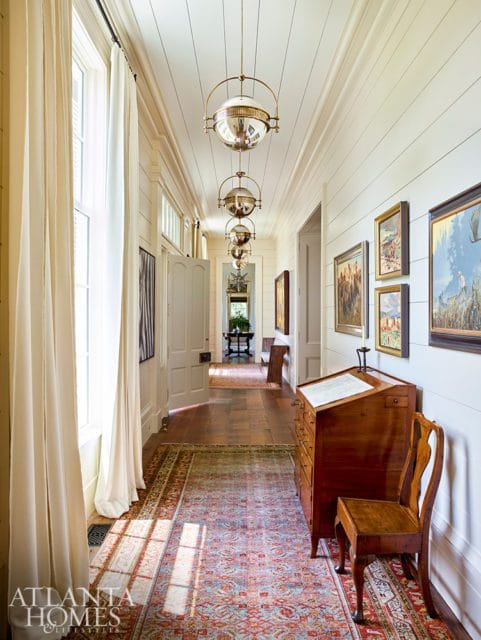 Hallway with shiplap walls, hardwood floors, and a long runner rug. Features a wooden desk with a chair, several framed paintings, and a series of globe pendant lights hanging from the ceiling.