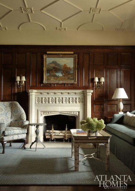 Living room with dark wood paneled walls, a white carved stone fireplace, a landscape painting, and upholstered furniture.