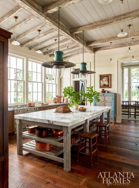 Farmhouse kitchen with marble island, exposed wooden beams on ceiling, and large windows. Copper pots and green pendant lights add rustic charm.