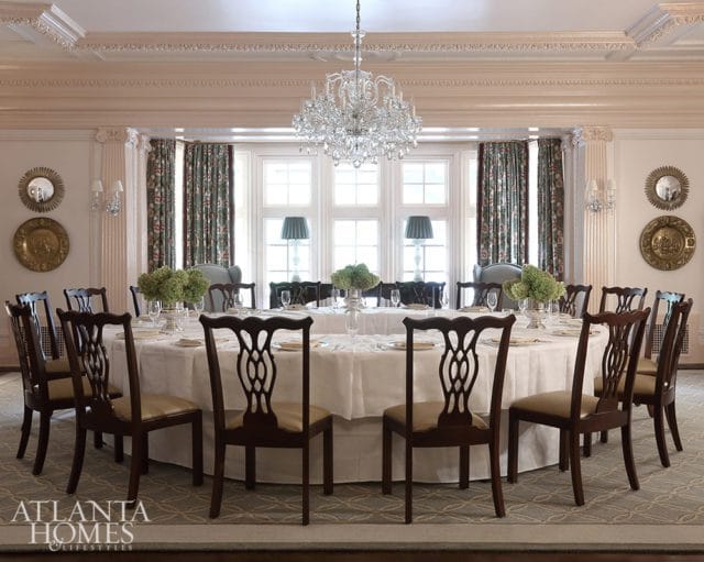 Formal dining room with a round table set for a meal, surrounded by dark wood chairs with upholstered seats. A crystal chandelier hangs above, and floral curtains adorn the windows.