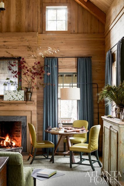 Living room with wood-paneled walls, a stone fireplace with a fire burning, blue curtains, and a round wooden table with four lime green chairs.