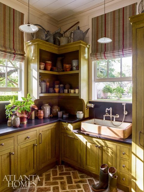 Yellow-green cabinetry with dark countertops and a farmhouse sink in a room with brick flooring and striped Roman shades.