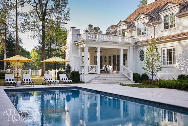 White brick house with a covered porch, balcony, and swimming pool. Four lounge chairs with yellow umbrellas are set up by the pool.