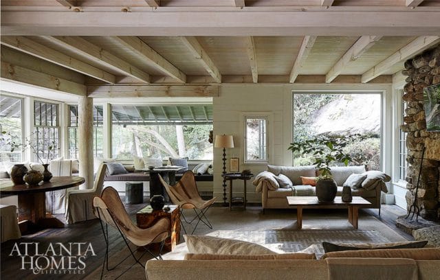 Living room with exposed wooden beams, stone fireplace, large windows overlooking greenery, and butterfly chairs.
