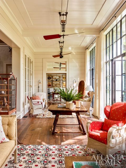 Sunroom with paneled walls, coffered ceiling, and large windows. Features wicker furniture, red upholstered chairs, a wooden table, and a patterned rug.