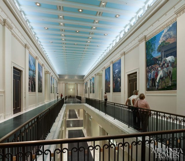 Hallway with a blue coffered ceiling, white walls adorned with large historical paintings, and dark railings overlooking a lower level.