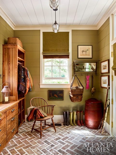 Mudroom with olive green walls, herringbone brick floor, wooden furniture including a tall cabinet and a chair, and various outdoor gear like boots and baskets.