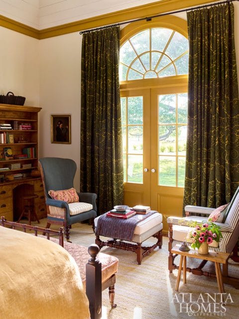 Bedroom with a large arched window and doors, dark patterned curtains, a wooden desk, armchair, ottoman, and striped accent chair.