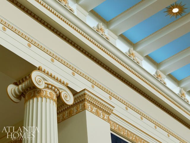 Interior detail of a room with ornate gold trim, a fluted column with a volute capital, and a blue coffered ceiling with a sunburst light fixture.