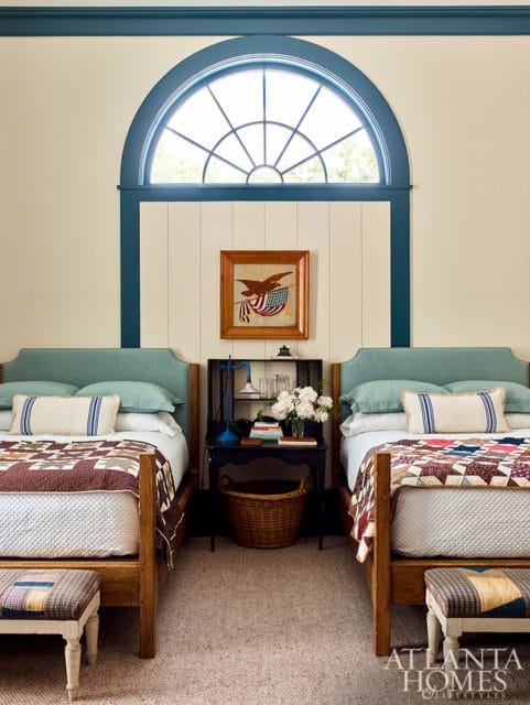 Bedroom with two twin beds featuring teal headboards, patchwork quilts, and a central nightstand with a basket. A large arched window with a fanlight is above a framed patriotic eagle artwork.
