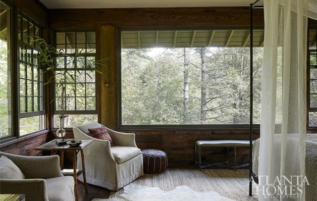 Cozy living area with wood-paneled walls, large windows overlooking a forest, comfortable armchairs, a leather pouf, and a bed with sheer curtains.