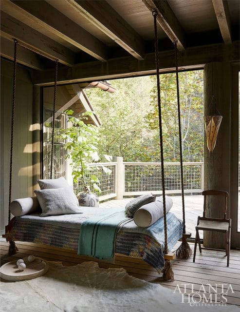 Porch with a hanging daybed covered in patterned textiles and light blue throw, overlooking a wooded area.
