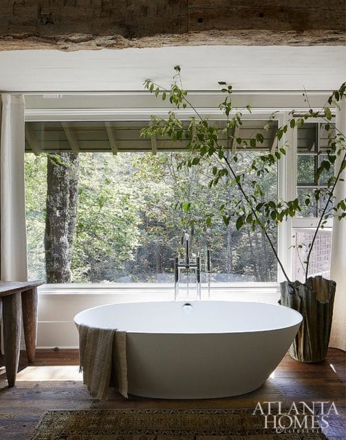 Freestanding white bathtub with chrome faucet against a large window overlooking a forest, with a rustic wooden table and a patterned rug.