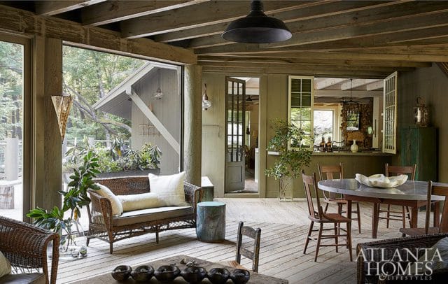 Covered porch with wicker seating, a dining table, and an open pass-through to a kitchen area. The walls are painted a muted green, and the ceiling features exposed wooden beams.