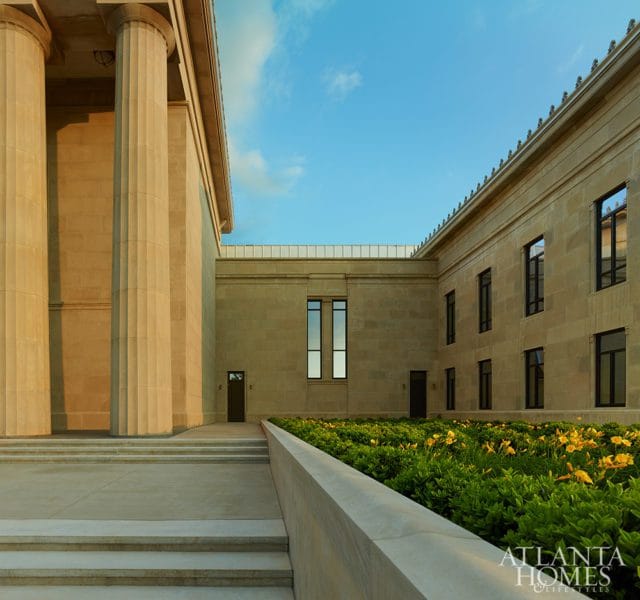 Classical building with large stone columns, stone facade, and a garden with yellow flowers under a blue sky. Atlanta Homes & Lifestyles logo in the lower right corner.