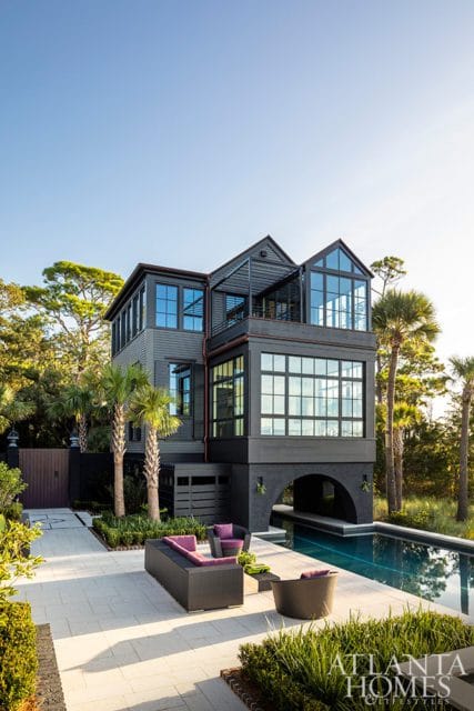 Dark gray modern house with multiple glass-enclosed levels, arched entryway, and swimming pool. Outdoor seating area on paved patio with palm trees and lush landscaping.