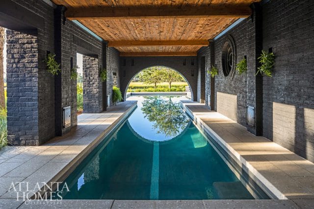 Long lap pool with dark brick walls, a wooden ceiling, and an arched opening revealing a tree-lined landscape.