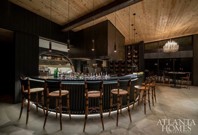 Modern bar area with a curved black counter, wooden stools, and a large stainless steel range hood. The ceiling is paneled wood, and a crystal chandelier hangs above a dining area.