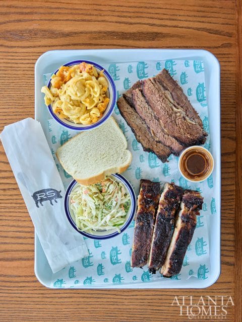 Barbecue platter with sliced brisket, ribs, macaroni and cheese, coleslaw, and a slice of white bread on a light blue tray with patterned paper.