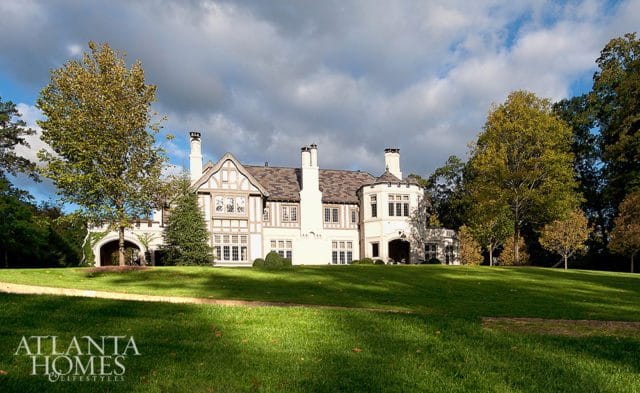 Tudor-style mansion with white stucco and dark timber framing, multiple chimneys, and arched entryway, set on a manicured lawn with mature trees under a cloudy sky.