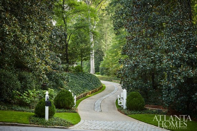 Winding driveway lined with lush green trees and manicured hedges, leading towards a large estate. White decorative fence posts and mailboxes flank the entrance.
