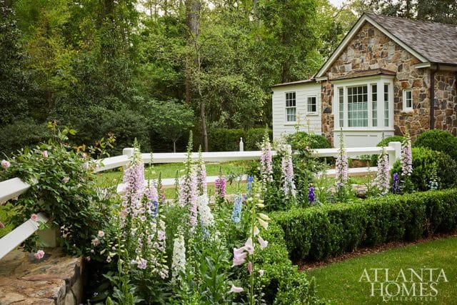 Stone cottage with white trim and bay window, surrounded by a garden with foxgloves, roses, and manicured hedges, with a white fence in the foreground.