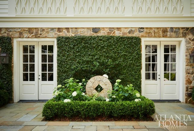 Two white French doors flank a dense green hedge wall, with hydrangeas and a stone millstone accent in the foreground. The house exterior features stone and white siding with decorative trim.