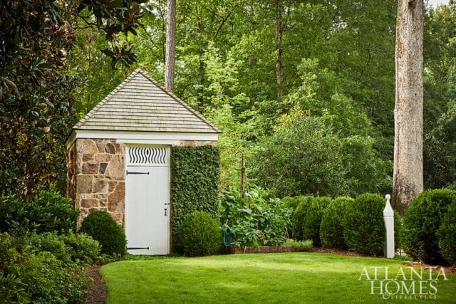 Stone garden shed with a shingled roof, white door, and ivy-covered wall, set in a lush green lawn with manicured hedges and mature trees.