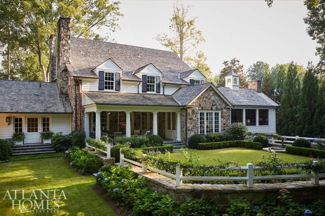 Stone and white clapboard house with a covered porch, dormer windows, and a cupola, surrounded by manicured gardens and mature trees.