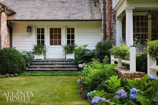 Stone and white clapboard house with a gray slate staircase leading to double white doors, flanked by lush green landscaping and blue hydrangeas.