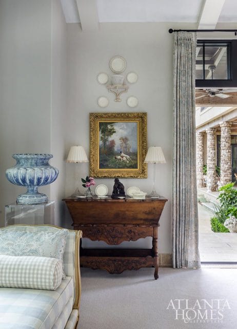 Living room with a blue and white ceramic urn on a lucite stand, a vintage wooden console table with a painting of dogs, and patterned curtains leading to an outdoor patio.