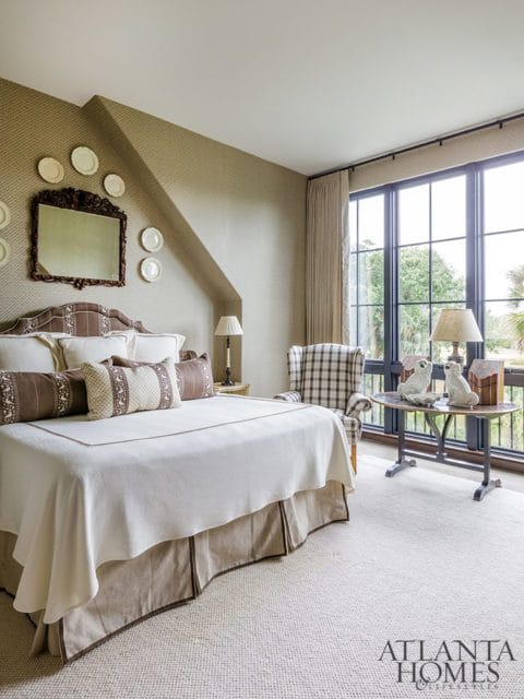 Bedroom with a large bed, patterned wallpaper, a decorative mirror, a plaid armchair, and a table with decorative objects by a large window.