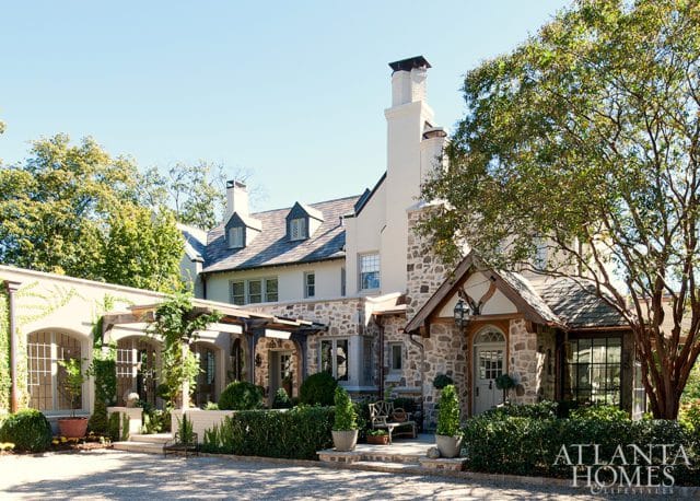 Tudor-style stone and stucco house with multiple gabled roofs, dormer windows, and a prominent white chimney. A covered walkway with arched windows and ivy leads to the main entrance.
