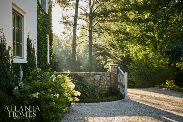 White brick house with ivy, stone wall, and open wooden gate leading to a gravel driveway surrounded by trees.