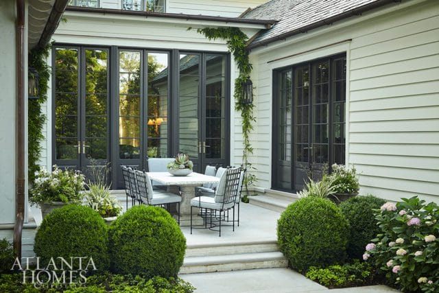 Patio with stone table and chairs set against a house with large dark-framed glass doors and windows. Lush greenery and boxwood shrubs surround the patio.