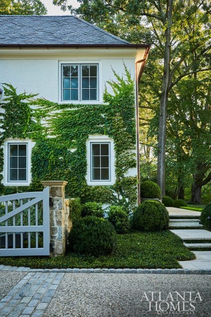 White stucco house with ivy climbing the walls, gray gate, stone pillar, and manicured landscaping.