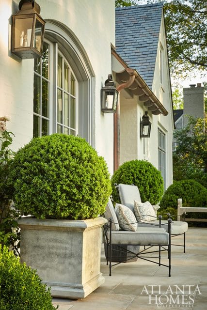 White stucco house exterior with arched window, slate roof, copper gutters, and black lanterns. Potted boxwood and outdoor seating on patio.