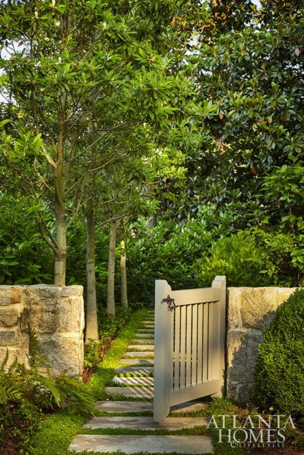 Stone pathway leading to a light gray wooden gate, flanked by stone walls and lush green trees and foliage.