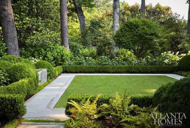 Formal garden with manicured boxwood hedges, a central lawn, concrete pathways, and lush surrounding trees and foliage.