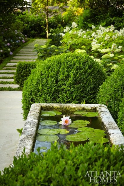 Stone water basin with lily pads and a single pink water lily, surrounded by manicured boxwood hedges and a stone pathway leading uphill through lush greenery.