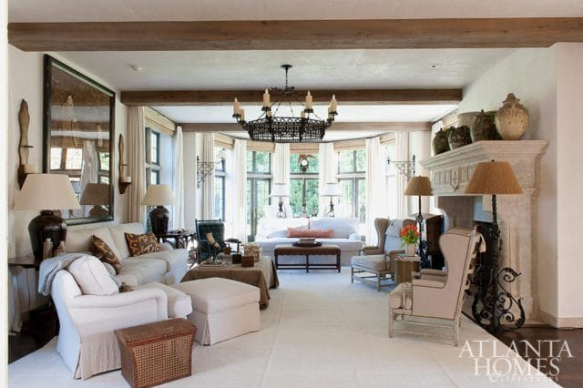 Living room with neutral-colored furniture, exposed wooden ceiling beams, a large stone fireplace, and a bay window overlooking greenery. Atlanta Homes & Lifestyles logo in the lower right corner.
