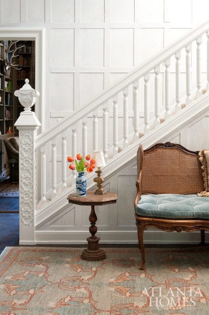 White paneled staircase with ornate newel post, a wicker bench with blue tufted cushion, and a small wooden table with a vase of tulips and a lamp.
