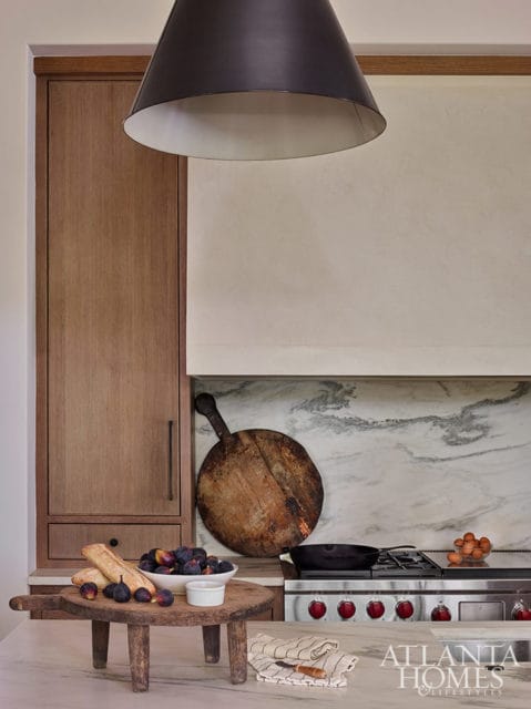 Kitchen with wood cabinetry, marble backsplash, stainless steel range, and a large black pendant light. A wooden cutting board with figs and bread sits on the counter.