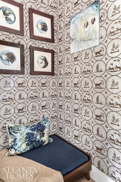 Corner of a room with patterned wallpaper featuring dogs and nature scenes, framed dog portraits, a blue cushion with a forest print, and a dark wood bench.