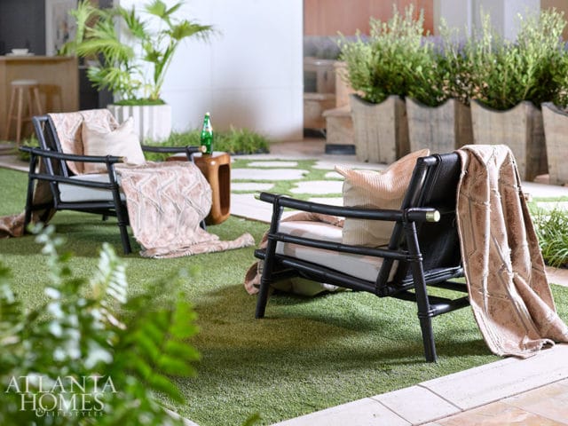 Two black rattan chairs with light cushions and textured throws sit on artificial grass, with potted plants and a bar area in the background.