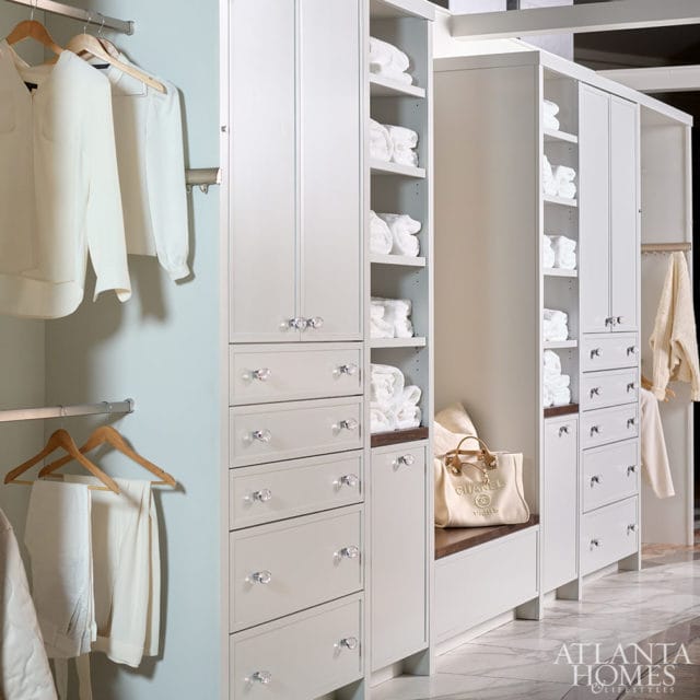 White closet with built-in drawers and shelves filled with neatly folded white towels. Clothes hang on racks to the left. A Chanel tote bag rests on a bench.
