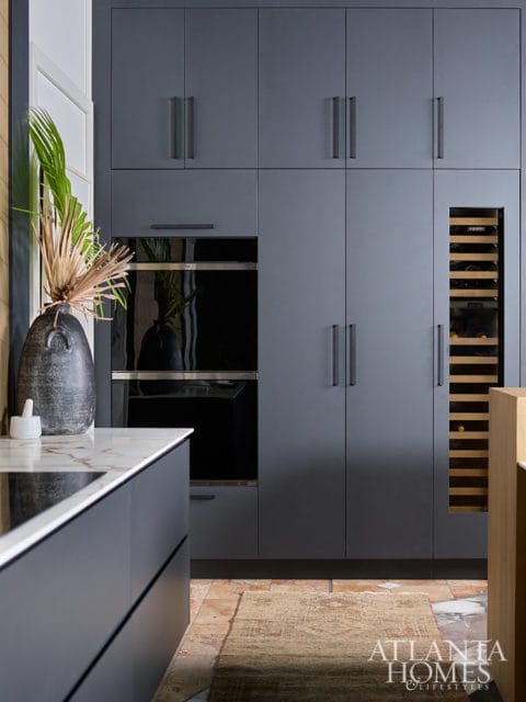 Modern gray kitchen with integrated ovens, wine fridge, and marble countertop. A decorative vase with dried foliage sits on the counter.
