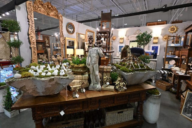 Antique store interior with ornate mirrors, statues, and decorative planters filled with moss and white objects.